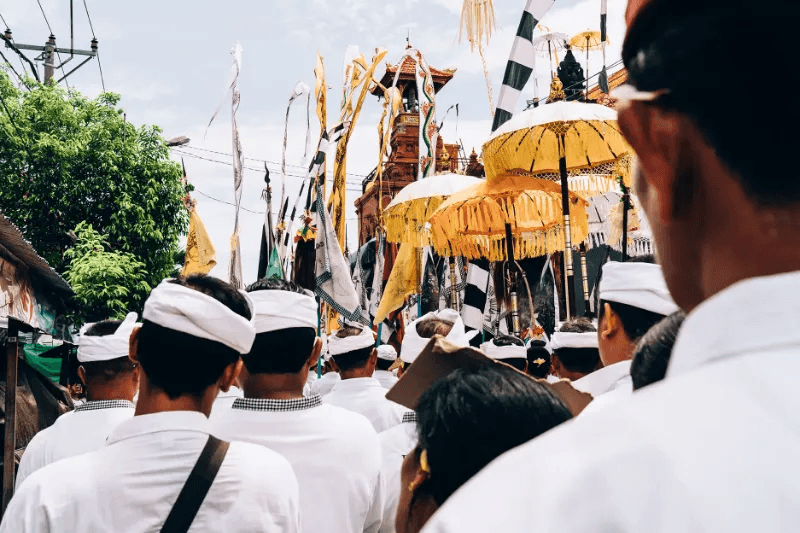 Back view of group of people in traditional clothes walking in decorated city with umbrellas and flags on crowded street in daytime in Bali
